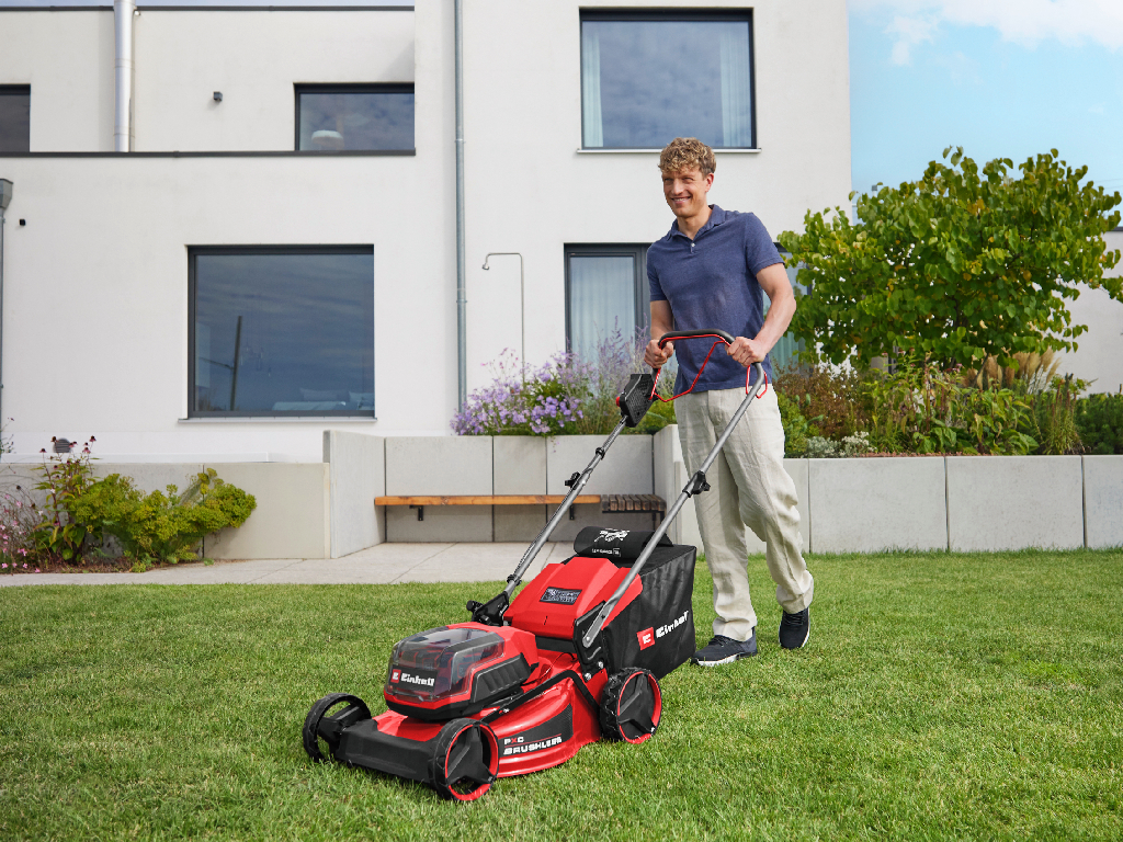 The picture shows a man wearing a red and black checked jacket using an Einhell cordless leaf blower to blow leaves into the air and away.